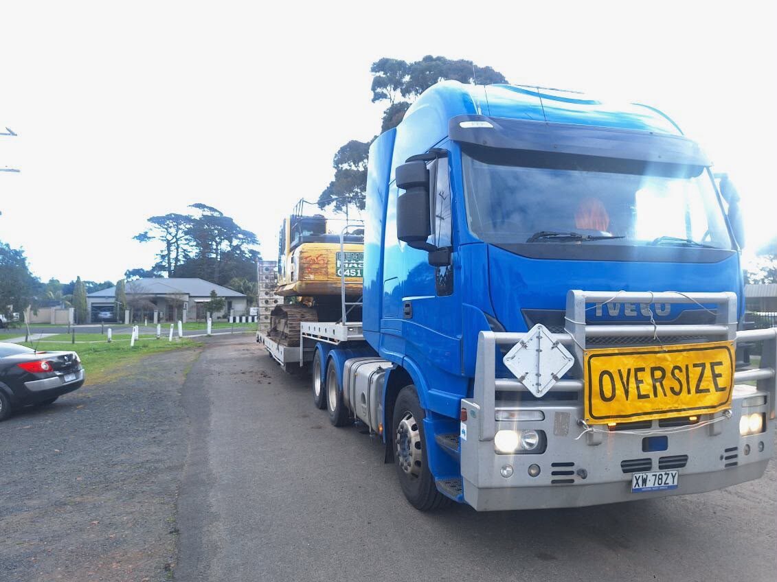 Blue prime mover with an oversize sign hauling a Demolition Masters excavator on a float trailer