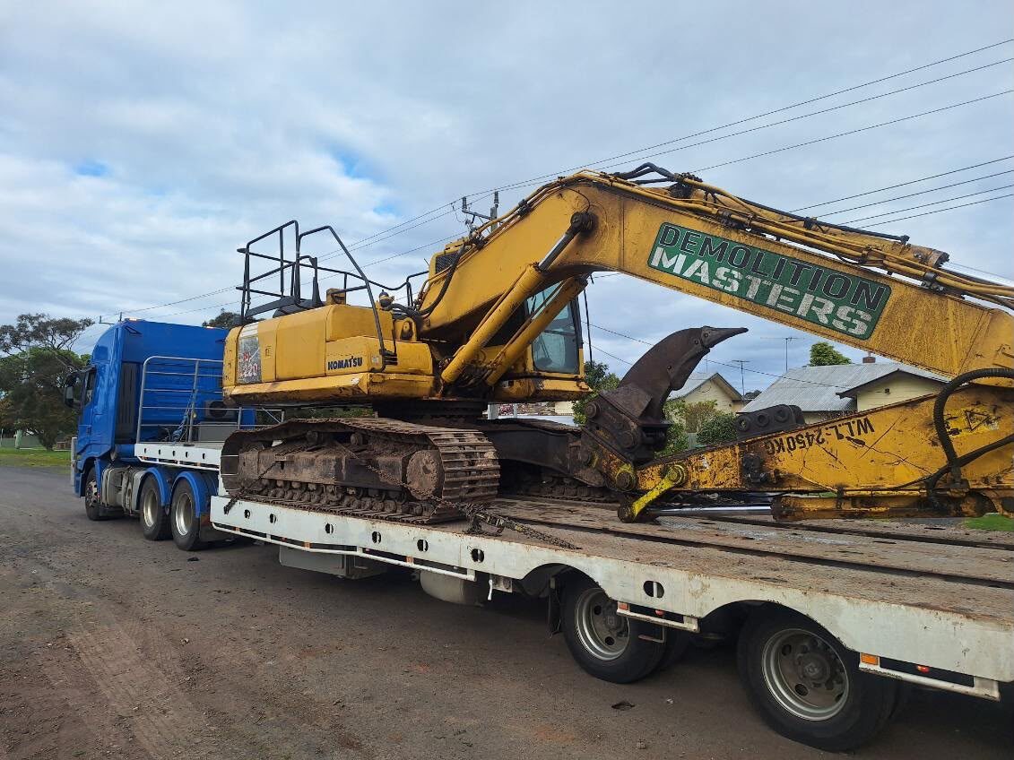 Yellow Demolition Masters Komatsu excavator with a pulveriser attachment loaded on a float trailer